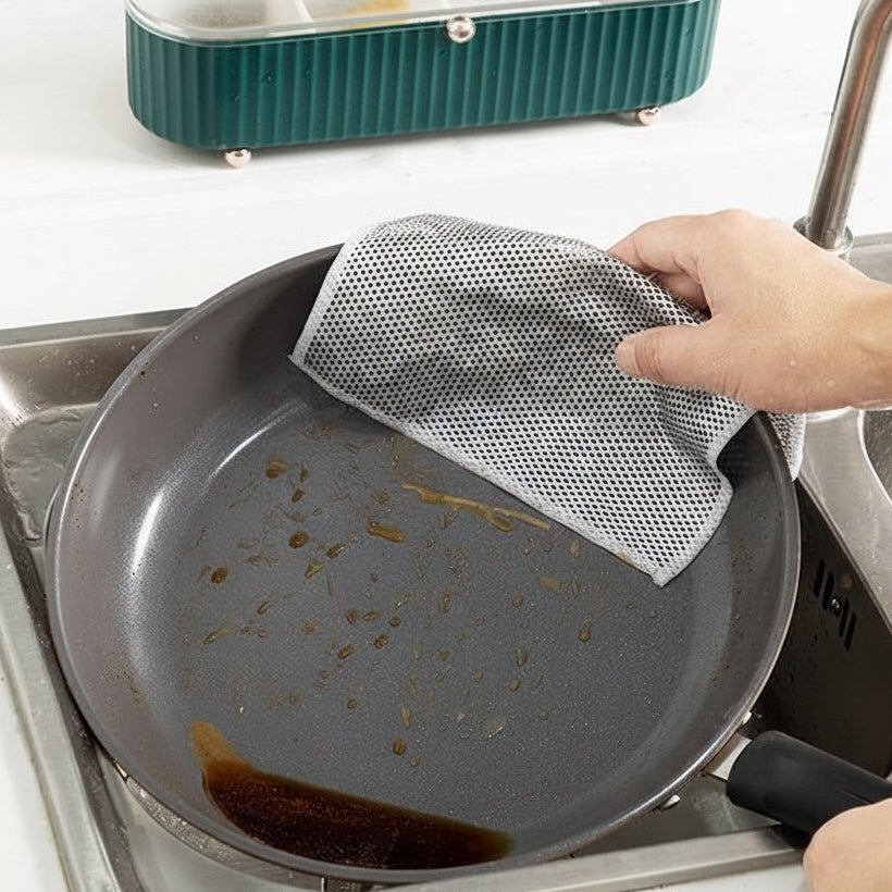 Person cleaning a greasy pan with a mesh scrubber in a kitchen sink.