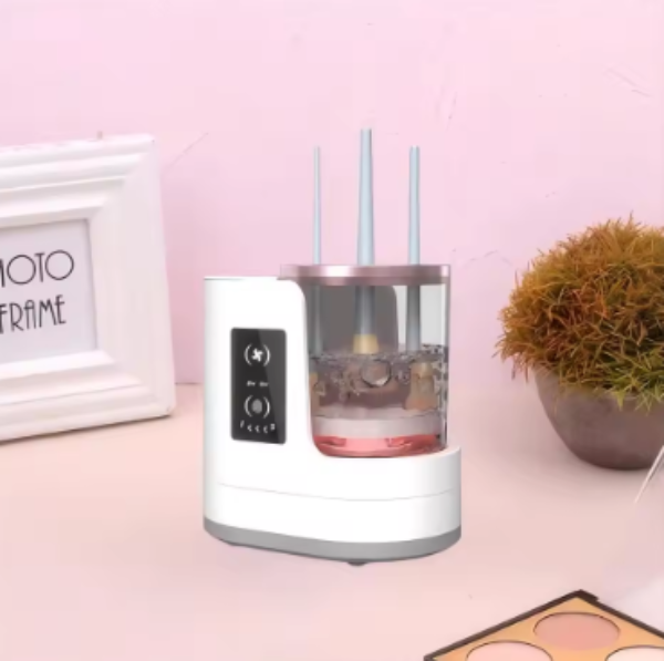 White Electric Makeup Brush Cleaner on a pink surface with decorative items.